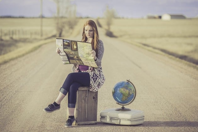 donna seduta su una valigia in mezzo ad una strada deserta guarda una cartina, accanto a lei un mappamondo poggiato su un altra valigia